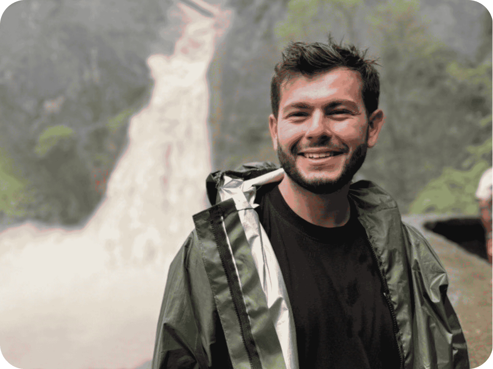 A medium close-up of a smiling man with a beard and short brown hair, wearing a dark rain jacket over a black t-shirt. He is outdoors with a blurred waterfall and rocky greenery in the background. The image has rounded corners and bright, natural lighting.