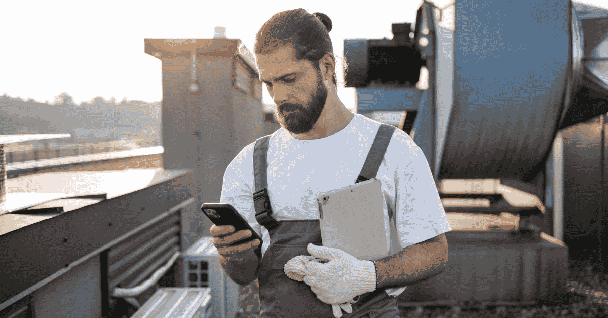 HVAC technician on a rooftop checking his phone and holding a tablet while on the job.