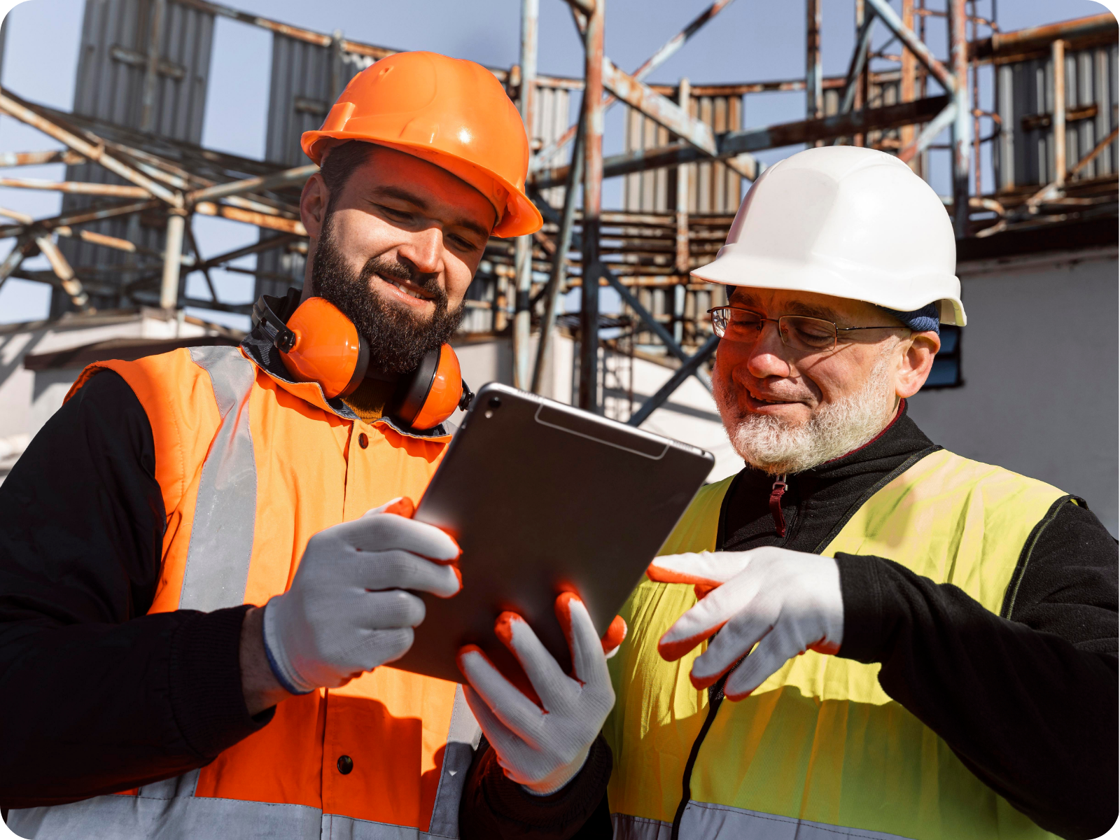 Two construction workers in hard hats and high-visibility vests reviewing project data on a tablet at a scaffolded job site.